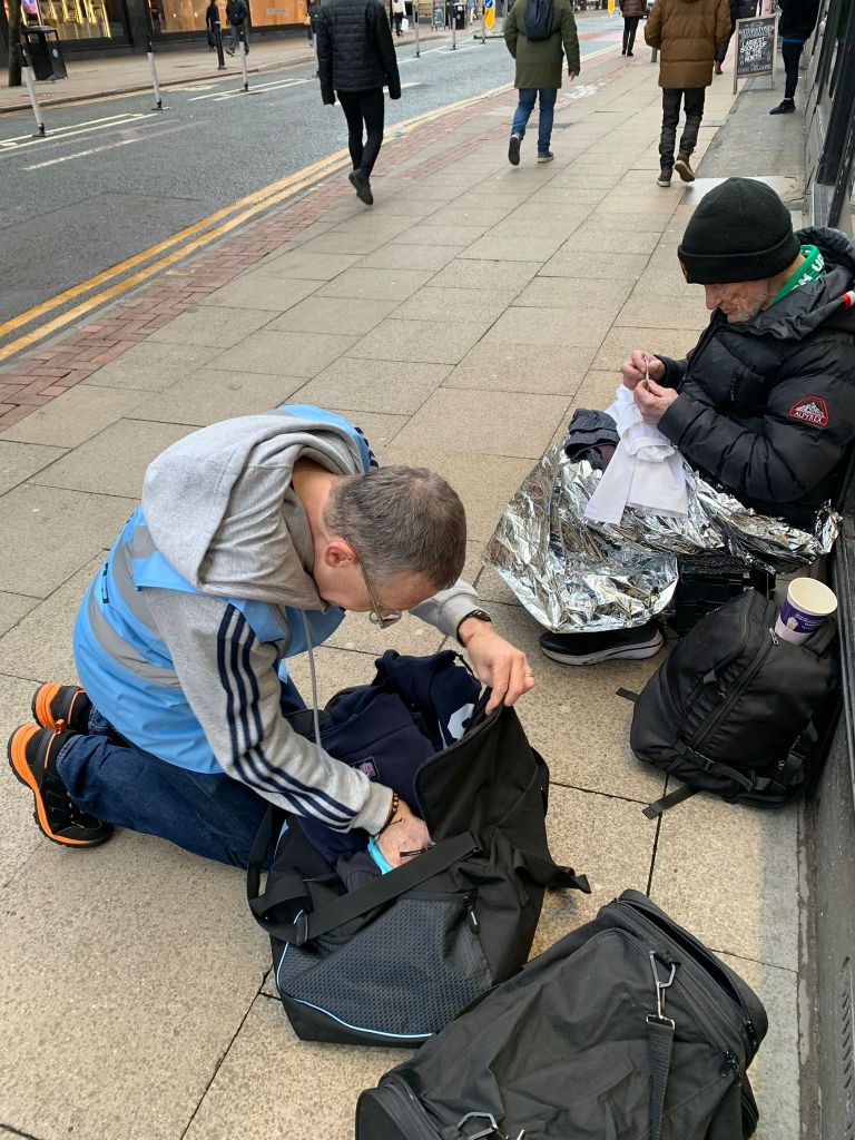 Two individuals on the street in Manchester, one kneeling and looking into a bag while the other sits nearby wrapped in a silver thermal blanket.