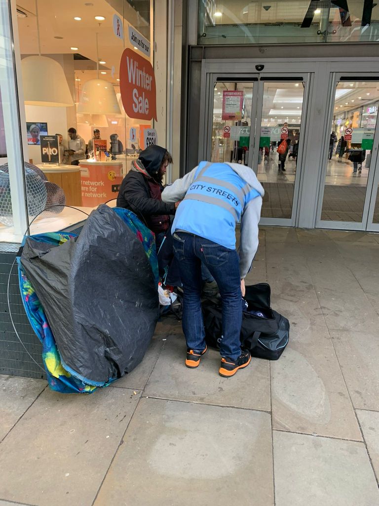 A City Streets volunteer assists a person experiencing homelessness outside a shop in Manchester, highlighting community support for those in need.